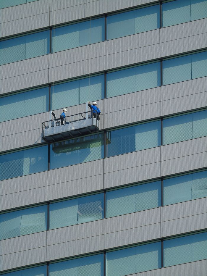 project-01-b Two workers clean windows on a modern glass building facade using a suspended platform.