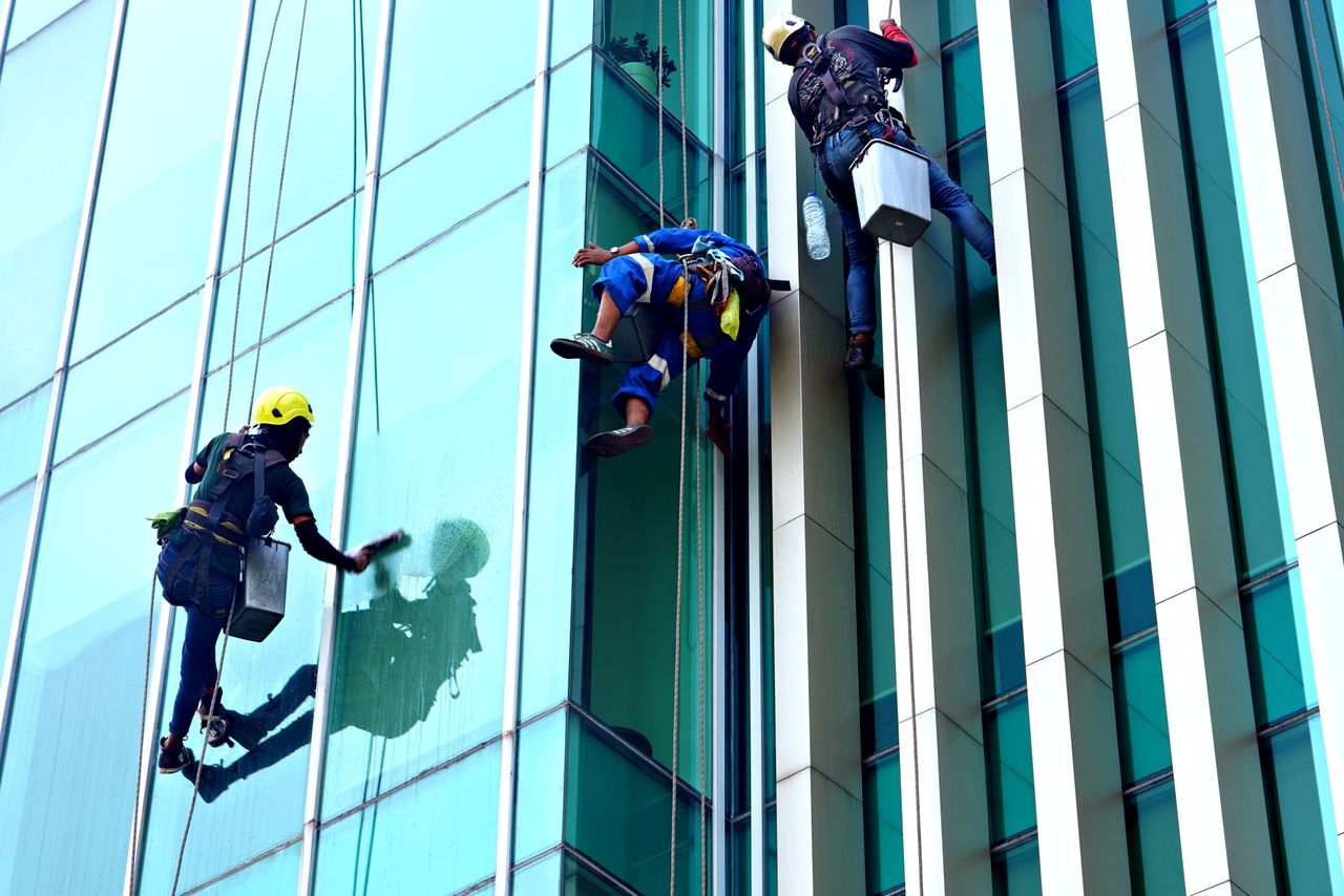 hero-img Three window cleaners suspended on ropes clean a modern glass building facade during the day.